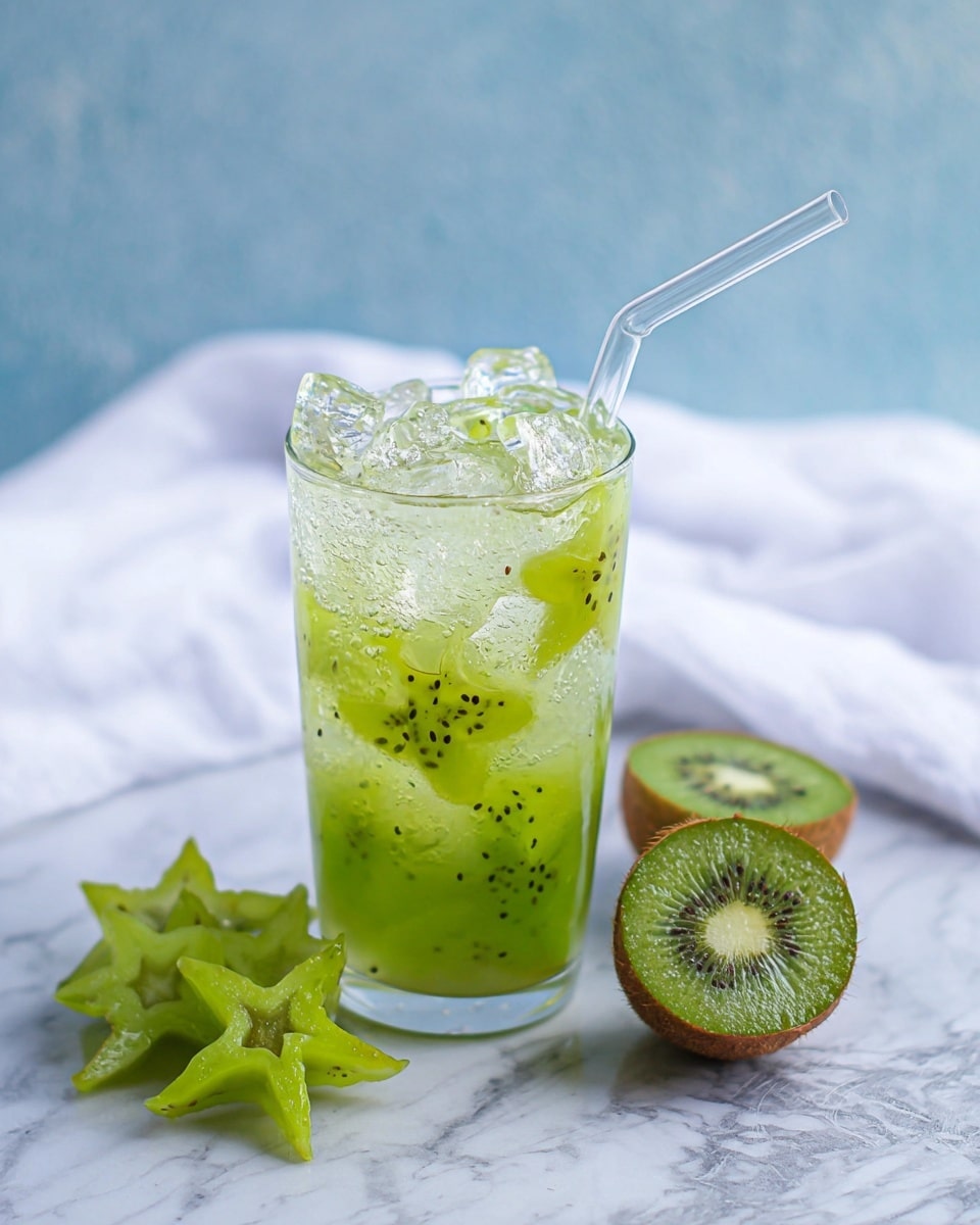 A tall clear glass filled with light green drink layered with many small ice cubes that have a shiny texture at the top, and slices of green starfruit shaped like stars floating inside the drink, with tiny black kiwi seeds visible in the liquid; a clear glass straw leans on the right side inside the glass. To the left side, green starfruit slices and one larger starfruit piece rest on a white marbled surface, while a cut kiwi fruit with green flesh and black seeds sits on the right. In the background, a soft white cloth is partially visible. Photo taken with an iphone --ar 4:5 --v 7