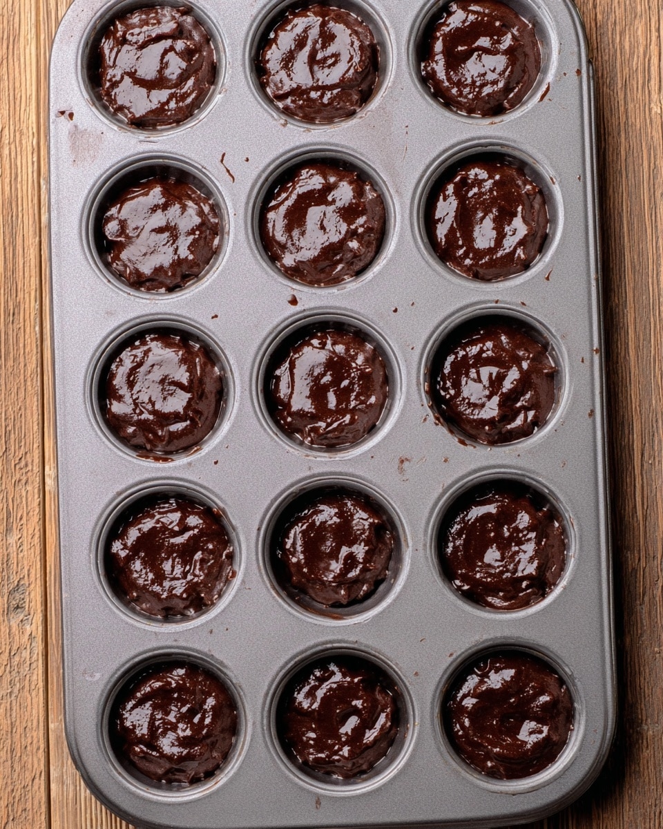 A gray metal muffin tray holds 24 round cavities, each filled with thick, dark brown chocolate batter. The batter has a glossy, slightly uneven texture with small swirls and soft peaks, filling each cavity more than halfway. The tray rests on a light brown wooden surface with visible grain lines. The metal surface shows some light oil spray and slight batter smudges around the edges of the cavities. photo taken with an iphone --ar 4:5 --v 7