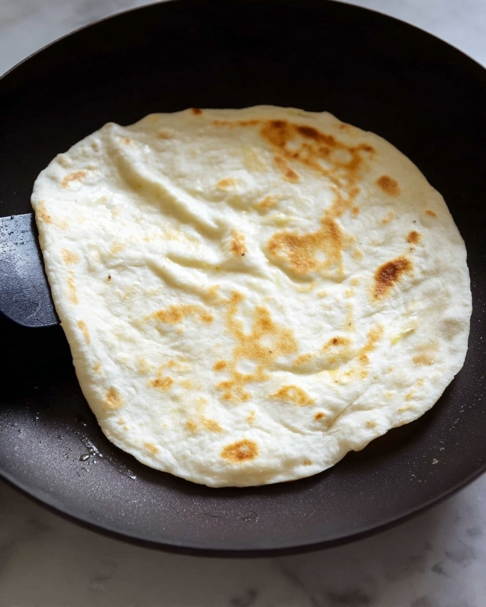 A single round flatbread is cooking in a dark pan, with light golden brown spots appearing on the white dough surface. The dough looks soft and slightly puffed in some places, showing gentle folds and airy texture. A black spatula is positioned to the left side, lifting or turning the flatbread. The background is a white marbled texture. photo taken with an iphone --ar 4:5 --v 7