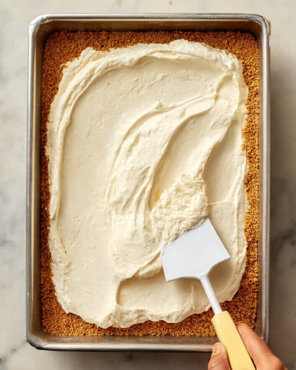 A rectangular metal pan holds two layers of dessert. The bottom layer is a crumbly, golden brown crust with a slightly rough texture. On top, there is a thick, creamy white layer being spread evenly with a white spatula that has a yellowish handle, which is held by a woman's hand. The background is a white marbled surface. Photo taken with an iphone --ar 4:5 --v 7