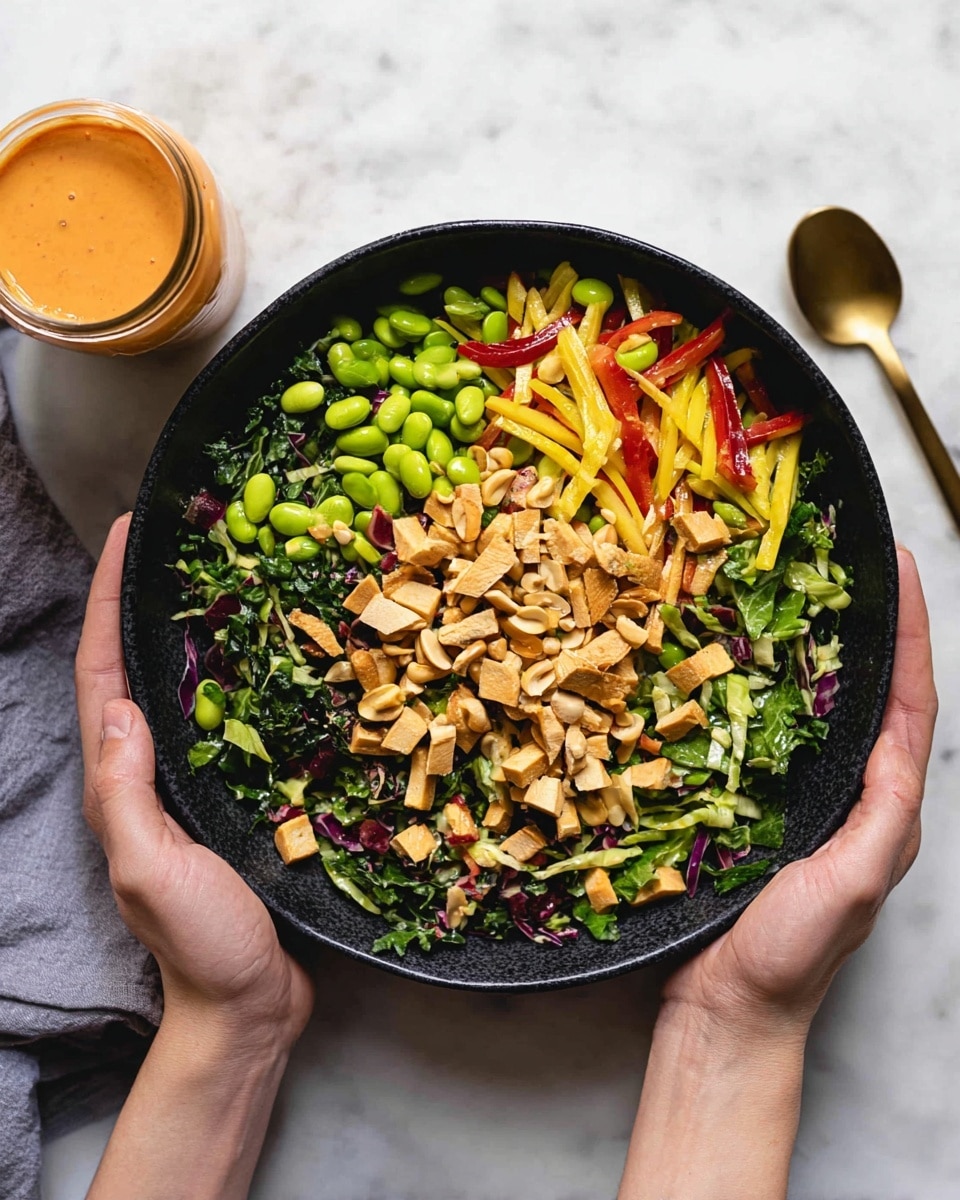 A close-up view of a large black bowl filled with a colorful salad, featuring multiple layers and textures: the bottom layer is dark green leafy greens mixed with light green shredded cabbage, the middle layer has thin yellow and red bell pepper strips along with green edamame beans, while scattered on top are light brown peanuts and sliced tofu pieces in a light golden hue. The bowl is held by two woman's hands against a white marbled surface, with a small glass jar of creamy orange dressing and a gold spoon placed in the background. Photo taken with an iphone --ar 4:5 --v 7