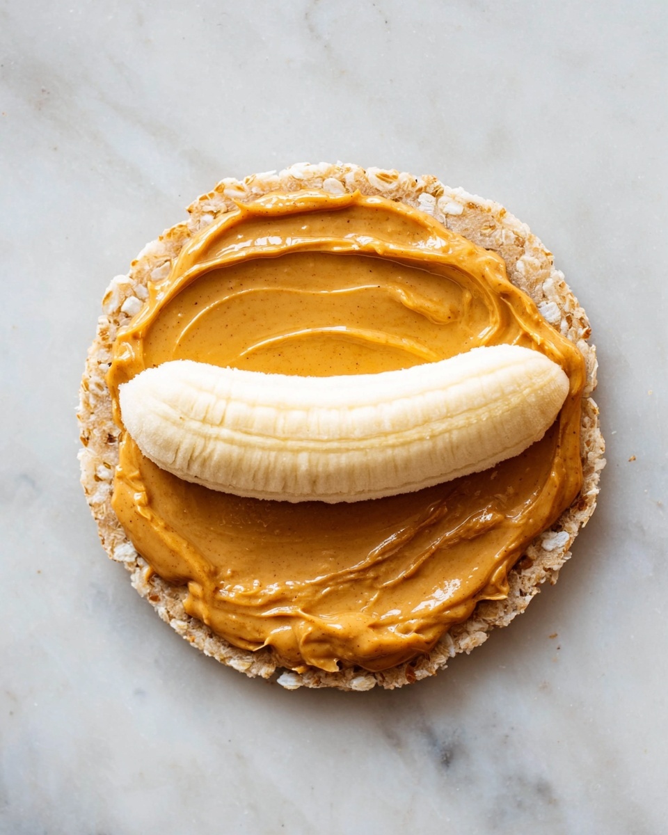 A round rice cake forms the base layer with a light beige color and a slightly rough texture. On top, there is a smooth layer of peanut butter spread evenly across the rice cake, showing a rich golden-brown color with a slight shine. Above the peanut butter, a whole peeled banana, light yellow with a smooth surface, is placed horizontally near the upper edge of the rice cake. The background is a white marbled surface. photo taken with an iphone --ar 4:5 --v 7