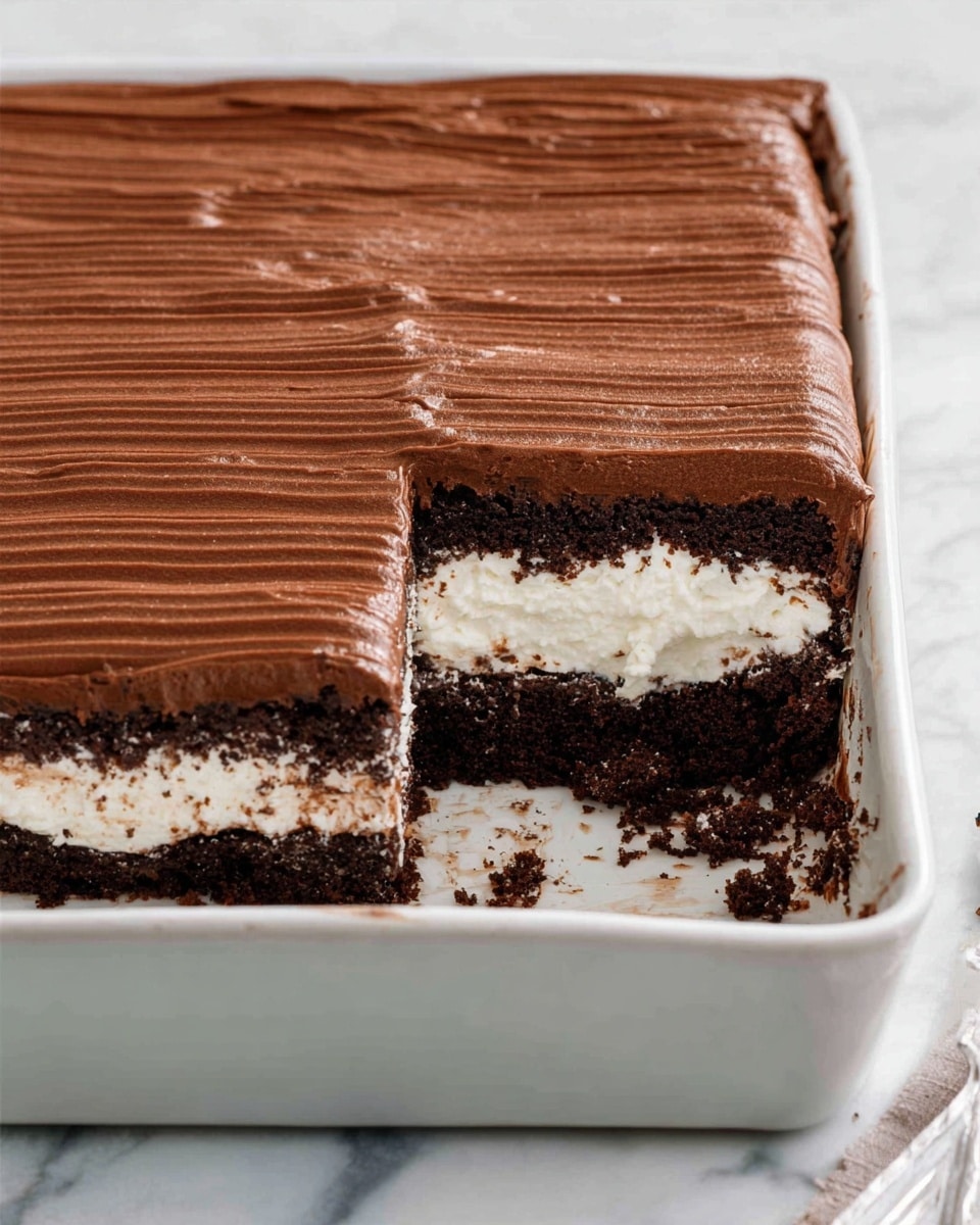 A close-up view of a three-layered dessert square on a white plate with a white marbled surface below. The bottom layer is a dark brown, soft chocolate cake with a crumbly texture. The middle layer is a thick, white creamy filling with bits of dark chocolate cake mixed inside, giving it a speckled look. The top layer is a rich, smooth chocolate frosting with fine horizontal ridges across its surface. Some small crumbs are scattered around the dessert on the plate. Photo taken with an iphone --ar 4:5 --v 7