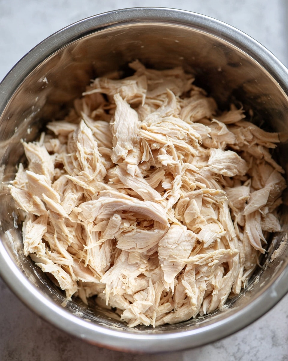 A close-up view of shredded cooked chicken in a shiny silver metal bowl, showing light beige chicken pieces with a fibrous texture piled up in the center, against the smooth, slightly glossy interior of the bowl, on a white marbled surface. photo taken with an iphone --ar 4:5 --v 7