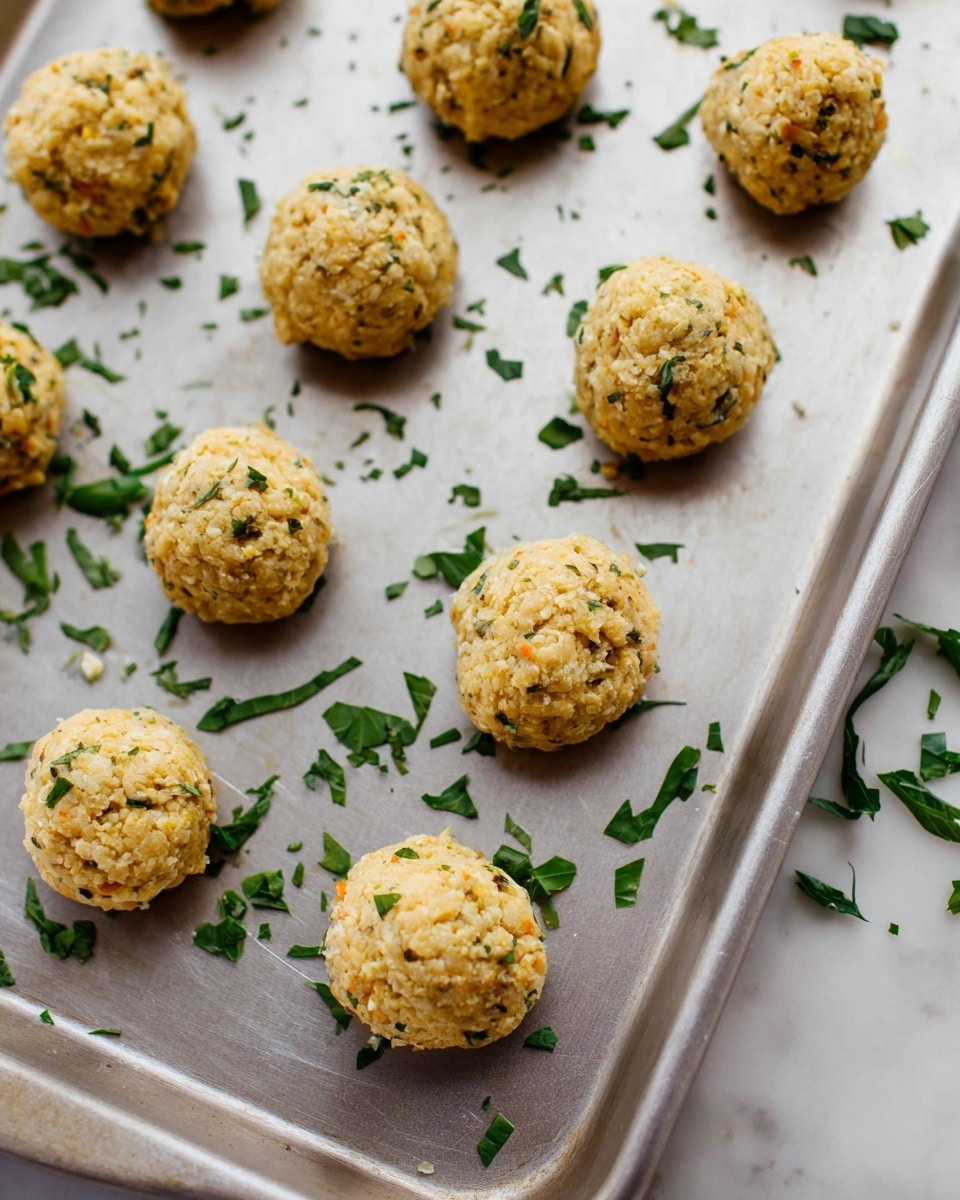 Several small round balls, light golden with hints of green herbs mixed in, are spread out evenly on a silver baking tray. The balls have a rough texture with small bits visible on the surface, and scattered fresh chopped green herbs are sprinkled around them on the tray. The setup sits on a white marbled surface. photo taken with an iphone --ar 4:5 --v 7