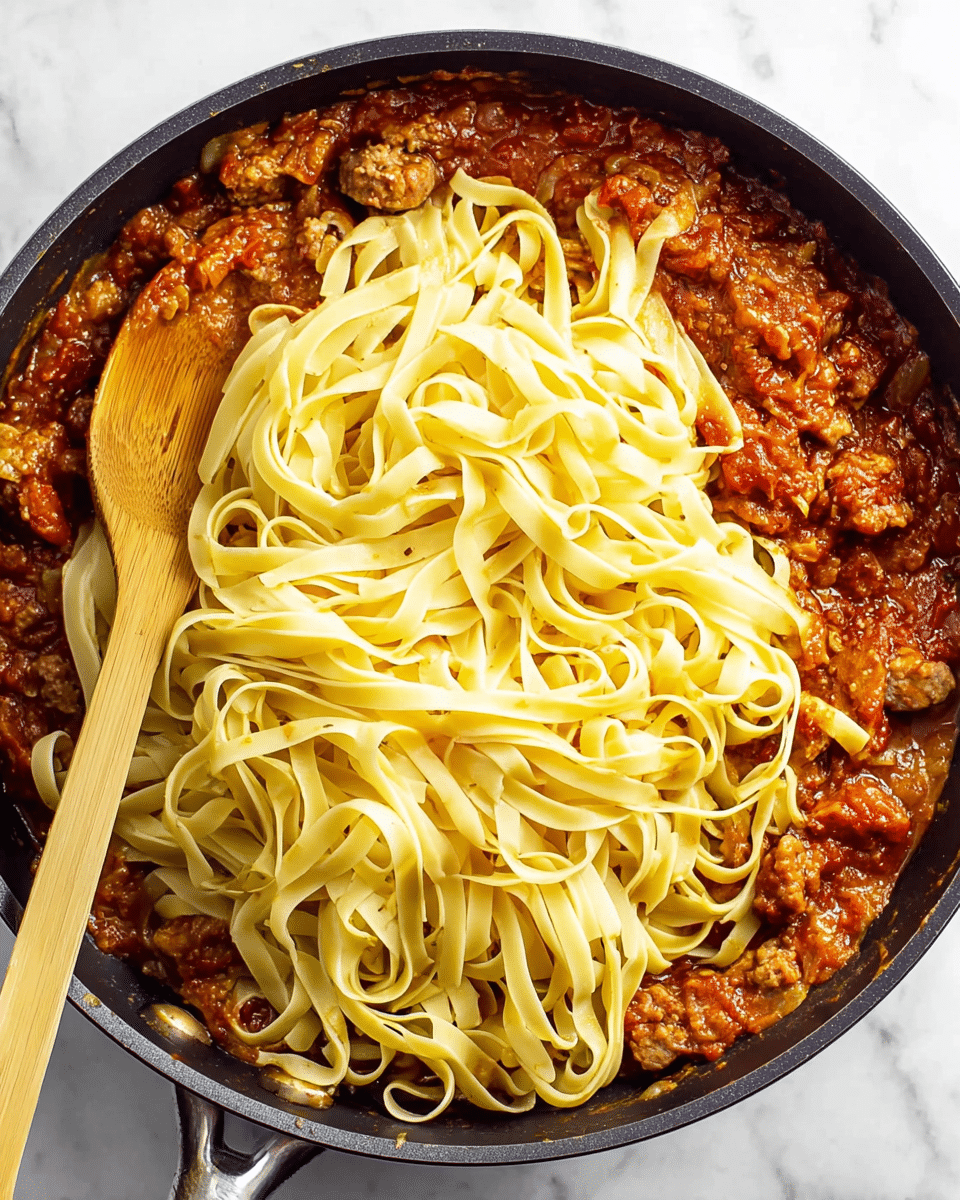 A large black pan filled with three visible layers: the bottom layer is a chunky, reddish-brown tomato sauce with bits of sausage, the middle layer has chunks of sausage partially covered by sauce, and the top layer consists of long, flat pale yellow noodles loosely placed over the sauce. A wooden spoon is partially submerged on the left side of the pan. The background is a white marbled texture. photo taken with an iphone --ar 4:5 --v 7