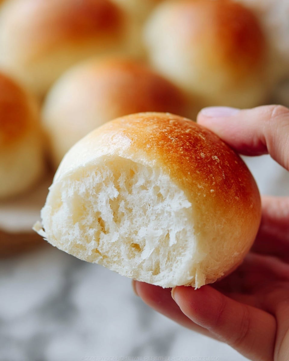 A close-up of a soft, round bread roll with a light golden-brown top crust and a fluffy, white inside showing a tender and airy texture. The roll is held delicately by a woman's hand with a blurred background showing similar bread rolls in soft focus on a white marbled surface. photo taken with an iphone --ar 4:5 --v 7