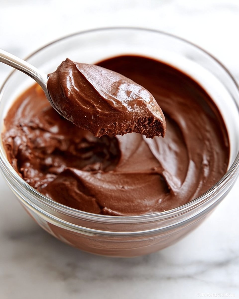 A close-up view of a clear glass bowl filled with a single thick layer of smooth, dark brown chocolate mousse with a glossy texture. A spoon is scooping some mousse from the bowl, showing its creamy and dense consistency. The bowl is placed on a white marbled surface. Photo taken with an iphone --ar 4:5 --v 7
