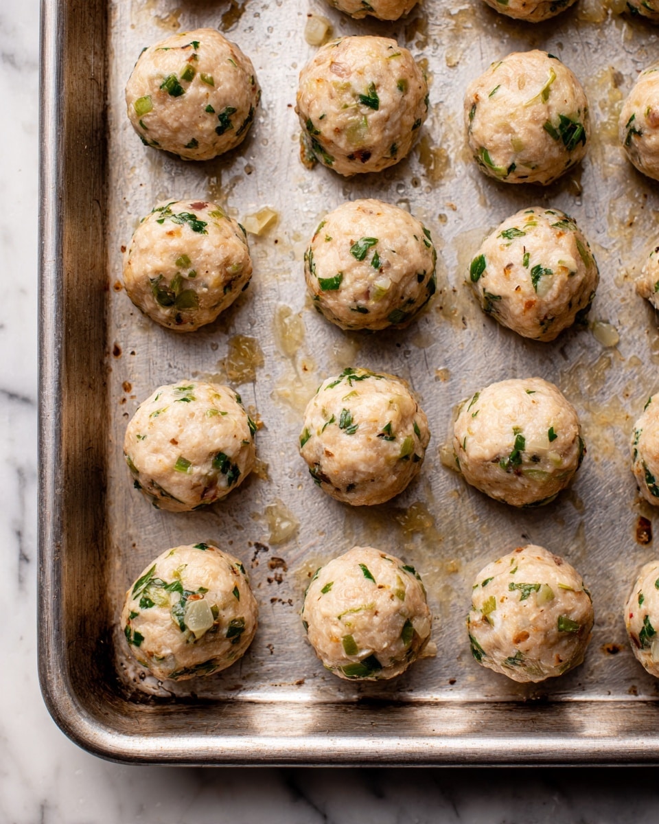 The image shows a metal baking tray filled with small, round, uncooked meatballs. Each meatball is light beige with visible bits of green herbs and small pieces of what looks like onion or garlic mixed throughout. The meatballs are arranged in neat rows, slightly spaced apart on the tray with some slight glistening areas from moisture. The tray has some spots of light browning and residue from previous use. The background is a white marbled texture photo taken with an iphone --ar 4:5 --v 7