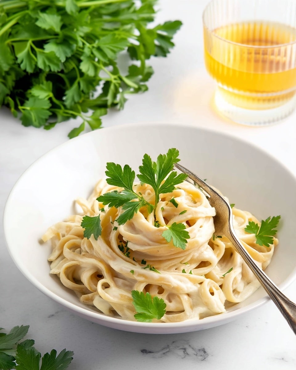 A white bowl holds a serving of creamy fettuccine pasta covered in a pale off-white sauce. The pasta strands are layered loosely in the bowl, with some gathered around a silver fork resting on the right side. Small green leaves of parsley and a single larger basil leaf are scattered on top, adding spots of bright green color. Light speckles of black pepper are sprinkled over the dish. The bowl sits on a white marbled surface with more green herb leaves and a glass with light golden liquid visible near the top left corner. photo taken with an iphone --ar 4:5 --v 7