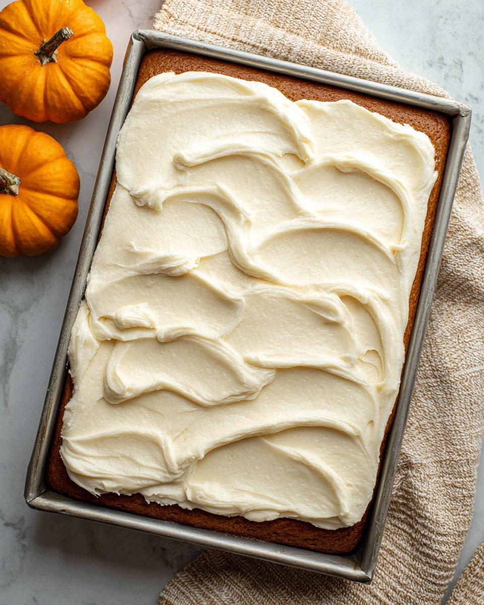 A close-up view of a rectangular cake in a metal baking pan, showing two layers: a thick, dark orange-brown cake base and a thick, creamy white frosting layer spread with swirls and soft peaks evenly covering the top. The edges of the frosting are slightly raised with a textured, smooth finish. The pan rests on a white marbled surface, with an orange mini pumpkin and beige textured cloth nearby. Photo taken with an iphone --ar 4:5 --v 7