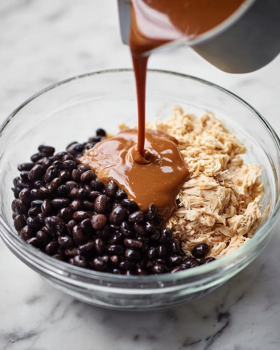 A clear glass bowl on a white marbled surface holds three layers side by side: shiny black beans on the left, light shredded chicken on the right, and a smooth brown sauce being poured from a grey container in the center. The sauce falls in a thick stream onto the beans, creating a wet dark pool that contrasts with the dry textures of the beans and chicken. photo taken with an iphone --ar 4:5 --v 7