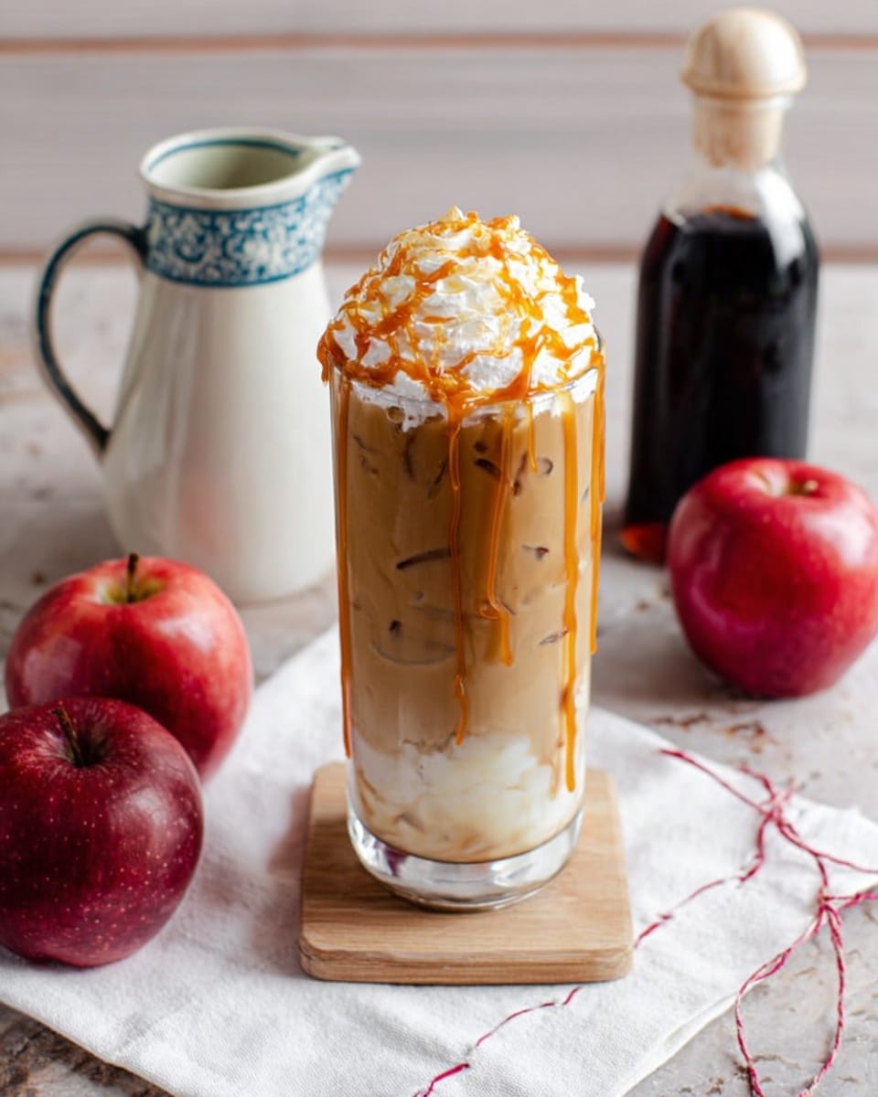 A tall clear glass filled with an iced coffee drink showing three visible layers: a light caramel brown liquid at the bottom with ice cubes, a thick fluffy white cream layer in the middle, and whipped cream topping with caramel sauce drizzled over the top and down the edges. The glass is placed on a small light wood board. Around it are two red apples, a white pitcher with a blue and beige rim, a dark liquid bottle with a white stopper, and a white napkin with red stitching, all set on a white marbled surface. Photo taken with an iphone --ar 4:5 --v 7