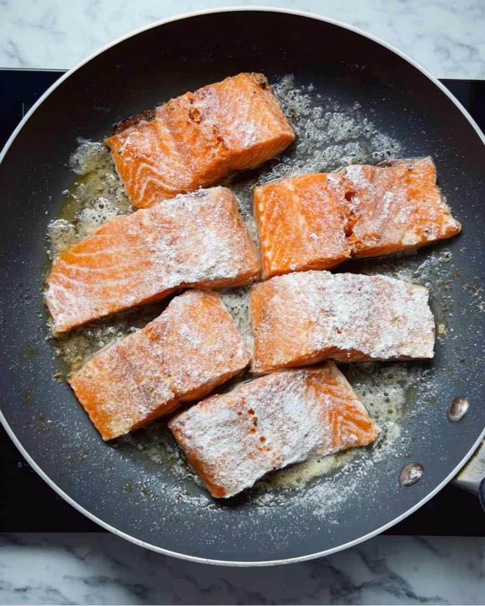 A gray non-stick frying pan holds four salmon fillets cooking inside it; each fillet is topped with a light white powder that looks like flour. The salmon pieces are a mix of light and dark pink with some orange tones, and the edges have a slightly crispy brown color. The fillets are arranged in a rough circle, with some gaps showing the pan’s smooth dark surface. The pan is sitting on a white marbled surface. photo taken with an iphone --ar 4:5 --v 7